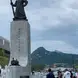 Statue of Admiral Yi Sun-sin in Gwanghwamun Square, Seoul, South Korea. The tall statue stands on a pedestal with Korean inscriptions, surrounded by a fountain and numerous visitors. The background features lush green mountains under a cloudy sky.