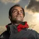 Picture of a man wearing a grey and red jacket, with a partly cloudy sky in the background