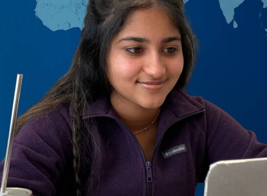 young woman smiling at computer with glass of water and a straw beside her and a virtual world behind her