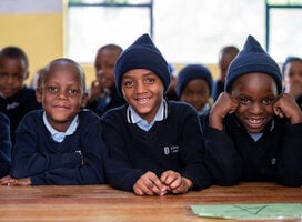 Three smiling boys in school uniforms, sitting together at a desk.