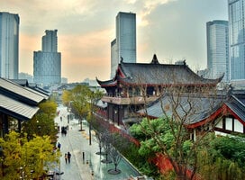 An image of Chengdu is shown. In the background, large city buildings can be seen, and in the foreground there are traditional Chinese buildings. On the left side of the image is a street lines with trees with people walking along it.