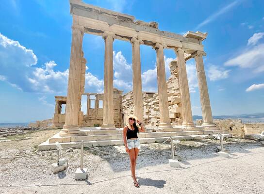 A young woman with long brown hair, wearing a black tank top and light denim shorts with a straw hat, stands in front of temple ruins with columns still standing.