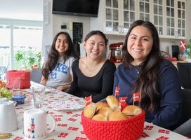 Three girls are sitting at a table and smiling. In front of them is a basket of baked good and Danish flags.