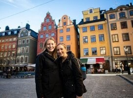 Two female students are standing in an old town square with colorful historical buildings behind them.
