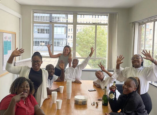 A group of people gathered around a table wave at the camera