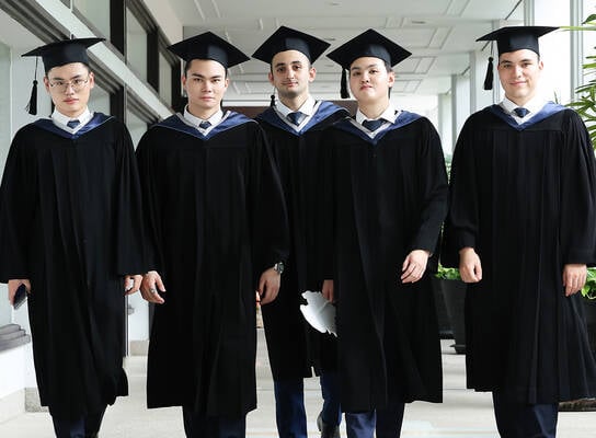 Five graduates in caps and gowns walk together down a hallway, facing forward and looking at the camera.