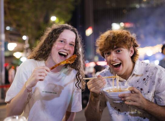 EF Gap Year students enjoying Japanese street food in Tokyo at night, laughing together while trying local dishes like ramen and skewers under the city lights.