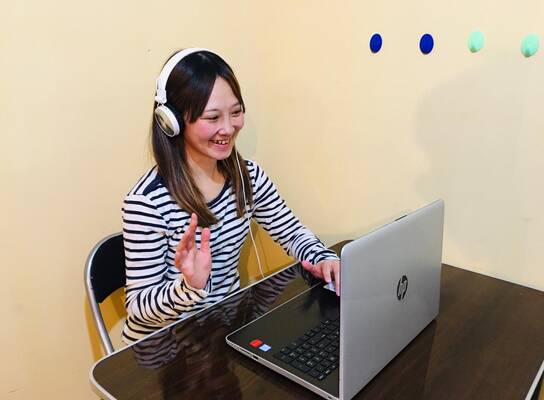 A Japanese teacher conducting an online 1-on-1 lesson with a student via Zoom. The teacher is smiling and explaining a lesson in Japanese using teaching materials on the screen.