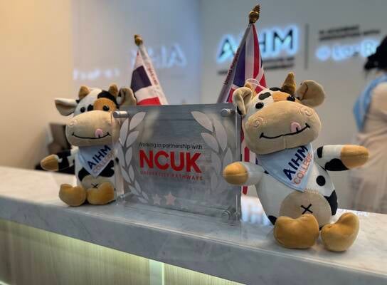 Two plush cow toys wearing AIHM College sashes sit on a reception desk next to a sign that reads, Working in partnership with NCUK University Pathways, with flags and logos in the background.