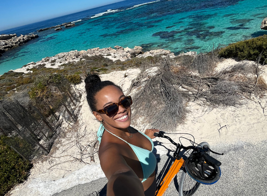 A girl on a bike taking a selfie with a blue sea behind her.
