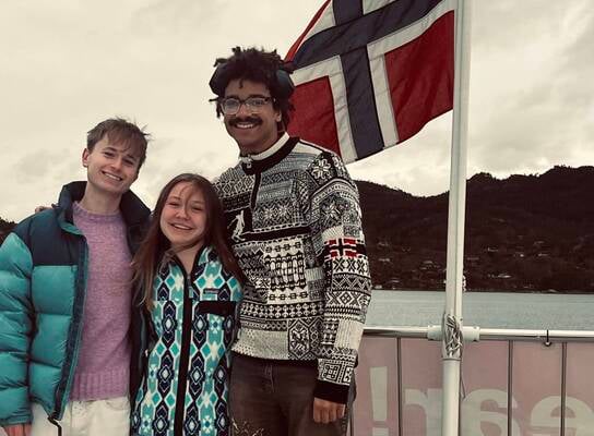 three college students stand smiling with the Norwegian flag waving in the wind
