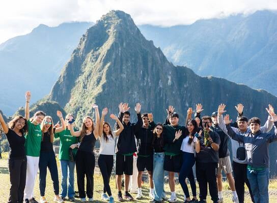 A group of teenagers standing on a stone terrace at Machu Picchu, waving their hands in the air with the ancient Incan ruins and Huayna Picchu mountain in the background.