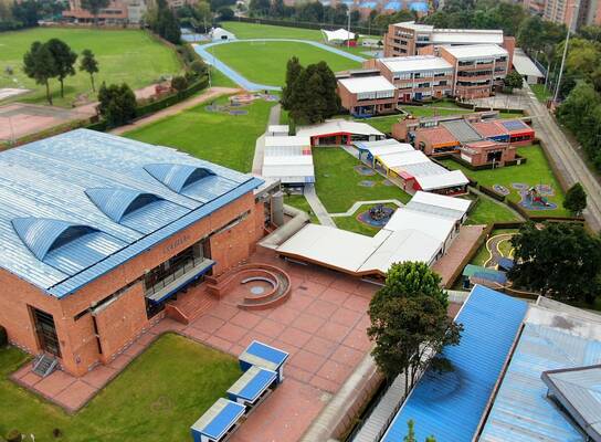 The image shows an aerial view of an educational institution with a spacious and well-organized campus, featuring brick buildings with blue and white roofs, well-maintained green areas, recreational spaces, and sports facilities such as an athletic track. Walkways and courtyards connect the different areas, and the surroundings combine nature with urban buildings, conveying a sense of order, openness, and institutional well-being.