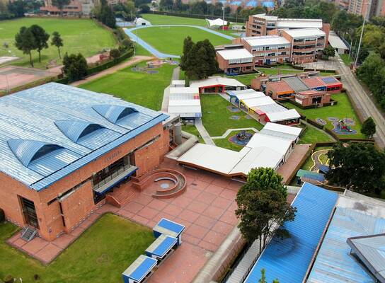 The image shows an aerial view of an educational institution with a spacious and well-organized campus, featuring brick buildings with blue and white roofs, well-maintained green areas, recreational spaces, and sports facilities such as an athletic track. Walkways and courtyards connect the different areas, and the surroundings combine nature with urban buildings, conveying a sense of order, openness, and institutional well-being.