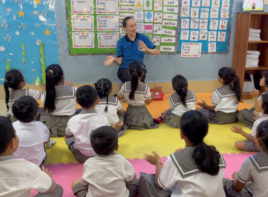 TEFL teacher sitting at the front of a classroom, engaging a group of young students in a lesson with hand gestures and visual aids on the wall.