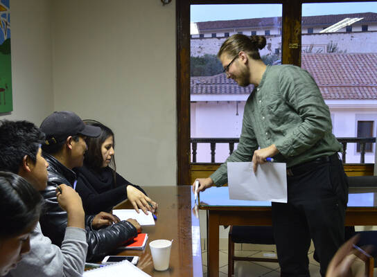 A man holding paper and a marker beside seated learners and a wooden table.