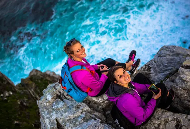 Two women sitting on a cliff by the ocean look up at and smile for the camera.