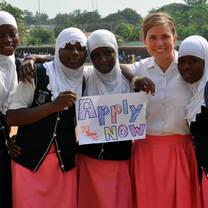 5 Women in white shirts and red skirts holding sign that says apply now in Ghana