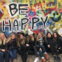seven girls sitting in front of the John Lennon Wall with the prominent phrase "BE HAPPY" behind them