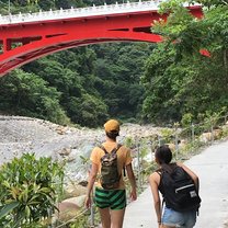 Friends walking together, with lots of plants around them and a tall, bright red bridge up ahead. Next to a small river bed