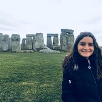 This photo shows a girl standing in front of Stonehenge. She is smiling and wearing a black coat, and it's cloudy behind her. The grass around Stonehenge is very green and contrasts nicely with the gray of the rocks. 