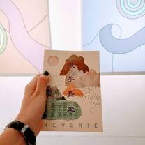 hand holding up a art show catalog which has mountains on it and the word " REVERIE ". Two abstract paintings are partially in view in the background.