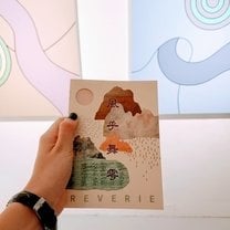 hand holding up a art show catalog which has mountains on it and the word " REVERIE ". Two abstract paintings are partially in view in the background.