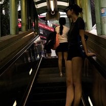 A girl wearing shorts, a t-shirt, and a baseball cap stands on an escalator, looking up and away from the camera.