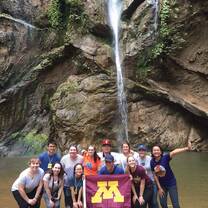 Large group of students smiling in front of a waterfall with the UofM flag. 