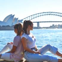 While on a trip to Sydney two best friends sit back to back looking at the Sydney Harbor Bridge and the Sydney Opera House.