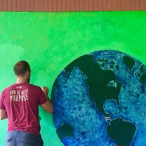 Travel With Purpose Man with back turned to the camera paints a large globe on the side of a building