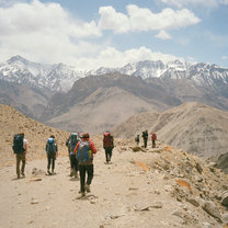 A landscape with snow-capped mountains in the background. Blue sky with clouds. Several hikers with backpacks walking away from the camera on a dirt trail.
