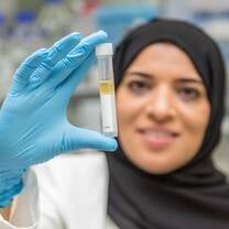 A chearful woman with a black headscarf and white labcoat holds a vile of liquid with hands protected in latex gloves in a modern laboratory