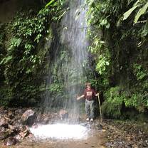 A man waves to the camera from behind falling water surrounded by lush green foliage