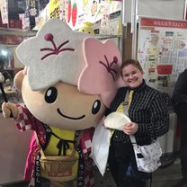 A Japanese Mascot with plum and sakura blossoms on its' head next to a person wearing a yellow tie and holding a pierogi 