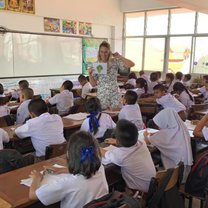 A teacher stands in front of a class of young Thai students, teaching them the word "broccoli" using flashcards. 
