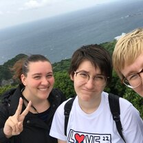Hiking excursion makes you tired. But still fun! The image is a horizontal picture of three girls from the chest up, looking at the camera. One makes a peace sign and smiles, another grimaces at the camera, and the last smiles at the camera as well. Behind them is a landscape with lush greenery, behind that the ocean, and a cloudy sky above them.