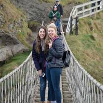 Two young women are standing in the middle of Carrick-a-Rede rope bridge, smiling at the camera with a green mountain view in the background.
