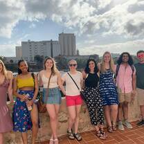 8 participants standing on an old balcony, smiling at the camera. The background is of a large high-rise business