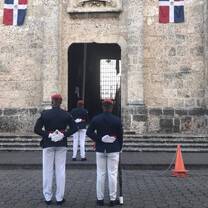 3 men in Dominican republic military uniforms are standing at guard in front of a pale building with two dominican republic flags hanging at each side