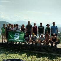 Travelors standing on a log across an overlook of Nepal while holding a green MSU flag in front of them. 