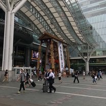 Hakata station. It is tall and there is a Yamakasa float in front of it. People walk around and the roof of the building is curved and made of glass.