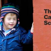 A little boy in blue coat and striped hat, smiles as he peeks through the door of a terra cotta colored play house. 