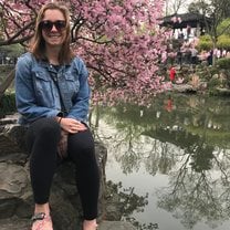 A young woman sitting on a rock with pink flowers behind her and a lake to the right