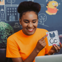 a woman in an orange shirt is smiling at a computer and pointing to a flashcard, which shows a picture of a cat and the letter C