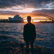 student gazing across the water at the sun setting behind the Sydney Opera House and Sydney Harbour Bridge