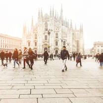 People walk across a stone plaza with a white gothic cathedral rising in the background