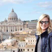 A smiling young blonde woman stands in front of the Vatican