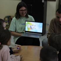 A group of children sit around a table, looking at two volunteer teachers. Once teacher holds a laptop computer with an image of a rocket ship on the screen.