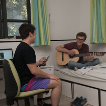 Two students seated, one of them plays a guitar